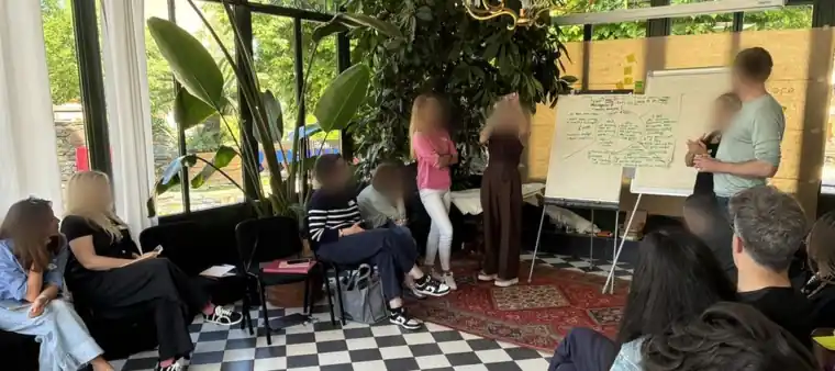 Groupe en cercle écoutant une restitution devant un paperboard, pendant une formation animée par RE-LIER — Group seated in a circle listening to a debrief at a flipchart during a RE-LIER training session